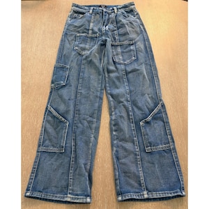 May include: Medium-wash blue denim jeans featuring a high-waisted design with a button and zipper fly closure. The pants include two front pockets at the hip, two lower cargo-style pockets on the thighs, and vertical seam detailing throughout.