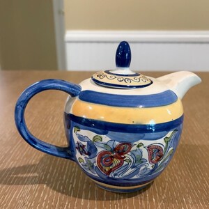 May include: A ceramic teapot featuring a hand-painted design with horizontal bands of blue and yellow. The body is decorated with intricate floral motifs in red, blue, and green. It includes a matching lid with a blue finial and a curved handle.