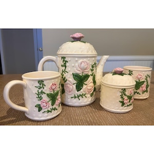 Op de afbeelding: Een crèmekleurige keramische theeset bestaande uit een theepot met deksel, een suikerpot met deksel en twee mokken. Elk stuk heeft een reliëf van witte stippen en is versierd met roze rozen en groene wijnranken met bladeren.