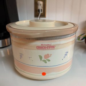 Può includere: Una pentola a cottura lenta in gres Rival Crock-Ette da 1 quarto con base e coperchio color crema. L'esterno presenta strisce orizzontali color pesca e blu, un piccolo motivo floreale e il testo Rival Crock-Ette 1 Qt. Stoneware Slow Cooker.