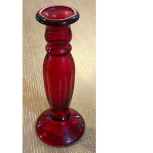 May include: A deep red glass candlestick holder featuring a smooth, rounded top rim, a slender neck with a subtle horizontal ring detail, and a bulbous, vertically ribbed body that tapers down to a wide, circular, flat base for stability.