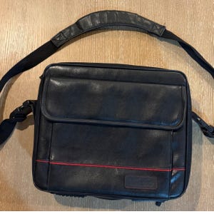 May include: A black rectangular messenger bag with a smooth finish, featuring a front flap, a thin horizontal red accent line, and a black adjustable shoulder strap with a padded section. The bag includes a small rectangular patch on the lower right.