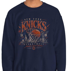 May include: Navy blue sweatshirt featuring a basketball graphic with the text "New York Knicks Basketball". The design includes a basketball going through a net, lightning bolts, and a cityscape silhouette. The sweatshirt has a crew neck.