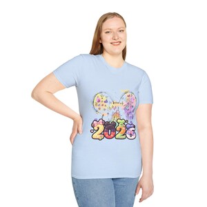 Puede incluir: Camiseta azul claro con un gráfico colorido que presenta el año "2026" y un diseño de orejas de Mickey Mouse. El gráfico incluye fuegos artificiales, corazones y un castillo. La camiseta es de cuello redondo.