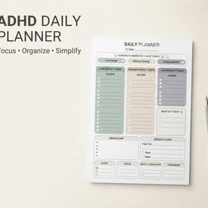 May include: A white daily planner sheet with sections for energy levels, tasks, brain dump, and mood. The text reads ADHD DAILY PLANNER Focus Organize Simplify. It features green, tan, and grey color-coded columns and a light green pen.