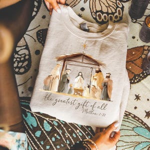 May include: A beige crewneck sweatshirt featuring a printed nativity scene illustration with a stable, figures, and a star. Below the scene, the text reads: the greatest gift of all Matthew 1:21 in a dark brown cursive and print font.
