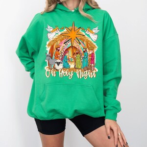 May include: A bright green hooded sweatshirt featuring a colorful graphic of a nativity scene with angels, the three wise men, a donkey, a sheep, and a star. The text "Oh Holy Night" is printed in a cursive font at the bottom of the graphic.