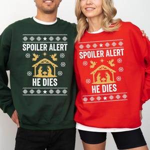 May include: A dark green sweatshirt and a bright red sweatshirt, both featuring a white and gold graphic print of a nativity scene with angels, snowflakes, and the text SPOILER ALERT HE DIES in a bold, sans-serif font across the chest area.