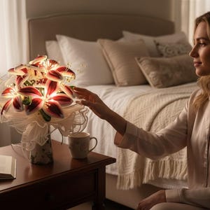 Op de afbeelding: Een boeket van verlichte lelies met rode en witte bloemblaadjes, gerangschikt in een decoratieve vaas. De bloemen staan op een donker houten nachtkastje naast een open boek en een mok met bloemenpatroon. De scène speelt zich af in een slaapkamer.