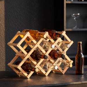 Puede incluir: Un botellero de madera natural que contiene varias botellas de vino. El botellero está hecho de listones de madera entrelazados y está diseñado para sostener las botellas en ángulo. Una sola botella marrón con tapa dorada está sobre la encimera.