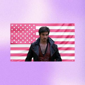 May include: A pink American flag with white stars and stripes. A man with dark hair and a black jacket with red accents is in front of the flag. The background is a gradient of purple.