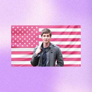 May include: A person wearing a dark gray jacket and a navy blue shirt stands in front of a pink American flag. The flag has white stars and pink stripes. The background is a light purple color.