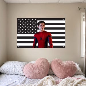 May include: A wall hanging featuring a black and white American flag design with a portrait of a person in a red and blue superhero suit. The image is mounted on a wall above a bed with heart-shaped pillows.