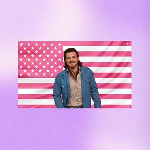 Puede incluir: Una bandera estadounidense rosa con estrellas y rayas blancas. Una persona sonríe frente a la bandera, vistiendo una chaqueta azul sobre una camisa a cuadros. El fondo es un degradado de púrpura.