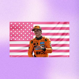 May include: A flag with a pink and white American flag design. A person wearing an orange racing suit and a cap stands in front of the flag. The suit has sponsor logos, and the cap has a McLaren logo.