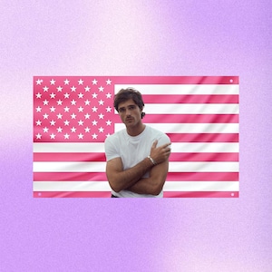 Puede incluir: Una persona con una camiseta blanca y una pulsera plateada, de pie frente a una bandera estadounidense rosa. La bandera tiene rayas rosas y estrellas blancas en la esquina superior izquierda. El fondo es un degradado de púrpura.