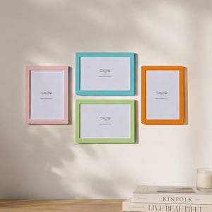 Op de afbeelding: Een set van vier rechthoekige fotolijsten in pastelroze, fel turquoise, limoengroen en oranje. Elke lijst bevat een witte inzet met de tekst CACTO in het zwart gedrukt. De lijsten zijn in een groep op een neutrale muur geplaatst.