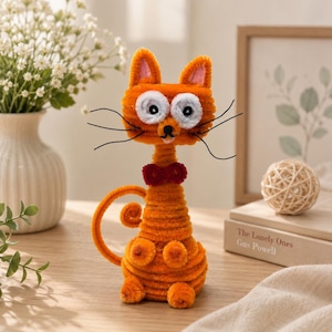 May include: A handcrafted orange cat figurine made from fuzzy pipe cleaners. The cat features large white circular eyes with black pupils, a small black nose, a red bow tie, and long black whiskers. Its tail is curled, and it has a textured body.