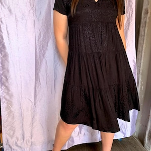 May include: A black tiered dress with short sleeves and a V-neck. The dress has a subtle pattern and falls to the mid-calf. The model is wearing black shoes. The background is a light purple curtain.