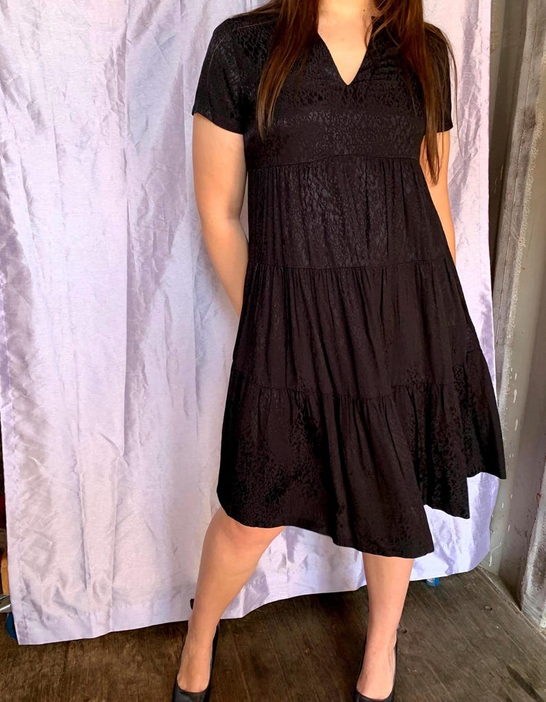 May include: A black tiered dress with short sleeves and a V-neck. The dress has a subtle pattern and falls to the mid-calf. The model is wearing black shoes. The background is a light purple curtain.