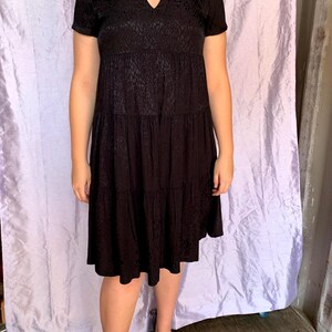 May include: Black short-sleeved dress with a V-neck and a tiered skirt. The dress features a subtle textured pattern. The model is wearing black flats. The dress is knee-length, suitable for various occasions.