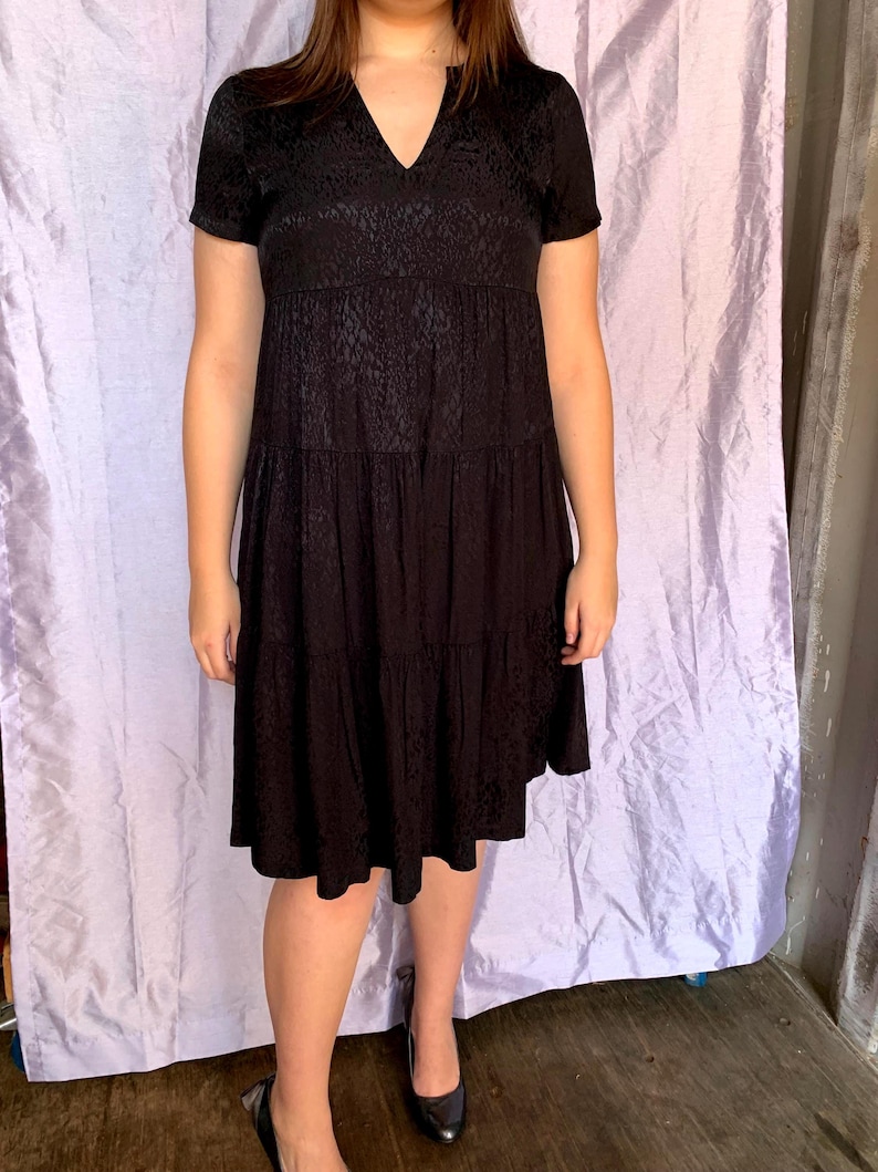 May include: Black short-sleeved dress with a V-neck and a tiered skirt. The dress features a subtle textured pattern. The model is wearing black flats. The dress is knee-length, suitable for various occasions.