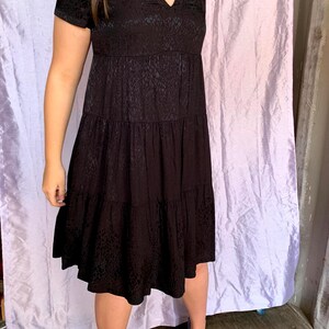 May include: A black, short-sleeved dress with a V-neck and a tiered skirt. The dress features a subtle textured pattern. The model is wearing black heels.