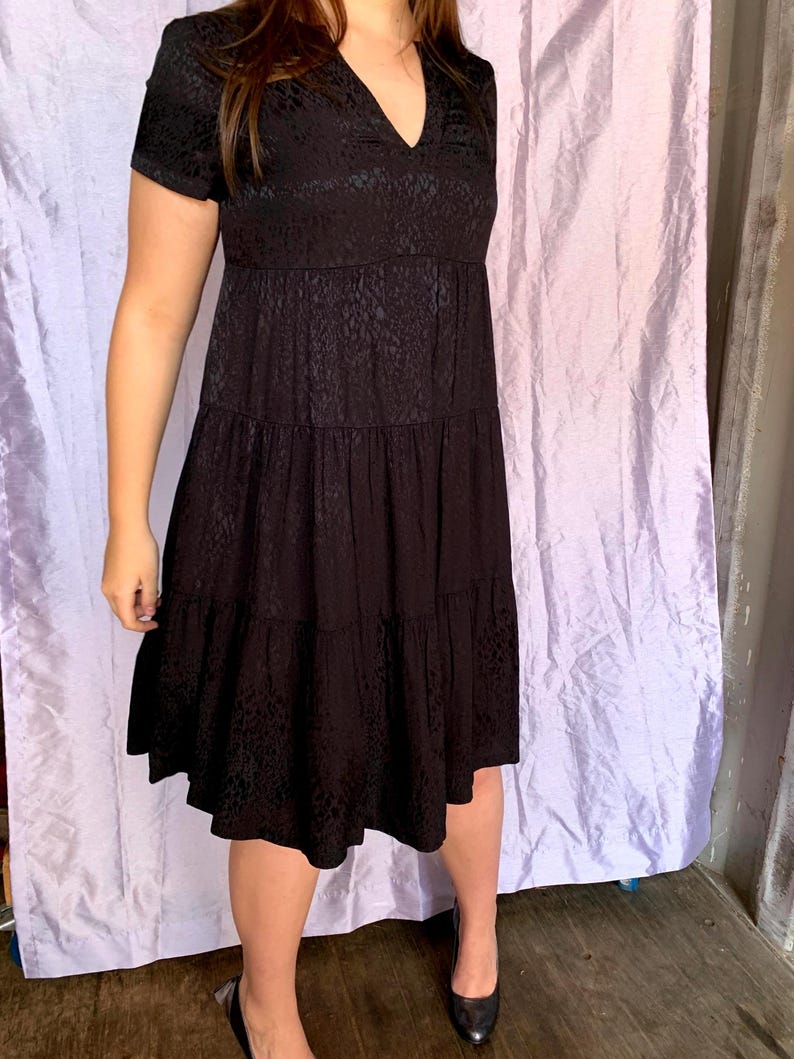 May include: A black, short-sleeved dress with a V-neck and a tiered skirt. The dress features a subtle textured pattern. The model is wearing black heels.