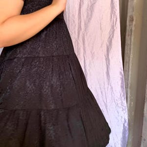 May include: A black dress with a textured pattern and tiered skirt. The dress has short sleeves and a flowing silhouette. The fabric appears to have a subtle sheen. The background includes a light purple curtain and a wooden surface.