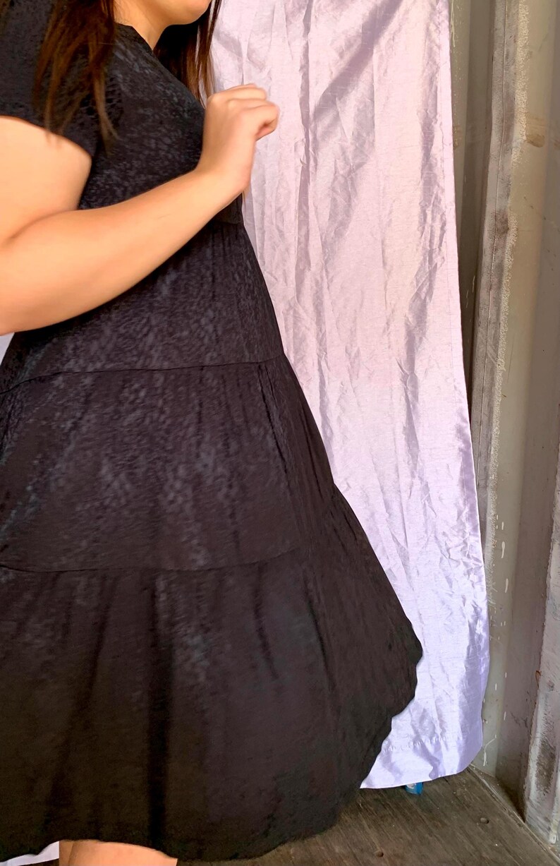 May include: A black dress with a textured pattern and tiered skirt. The dress has short sleeves and a flowing silhouette. The fabric appears to have a subtle sheen. The background includes a light purple curtain and a wooden surface.