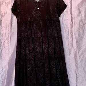 May include: A black, short-sleeved dress with a V-neck and a tiered skirt. The dress features a subtle, textured pattern. It hangs on a wooden hanger against a light purple backdrop.
