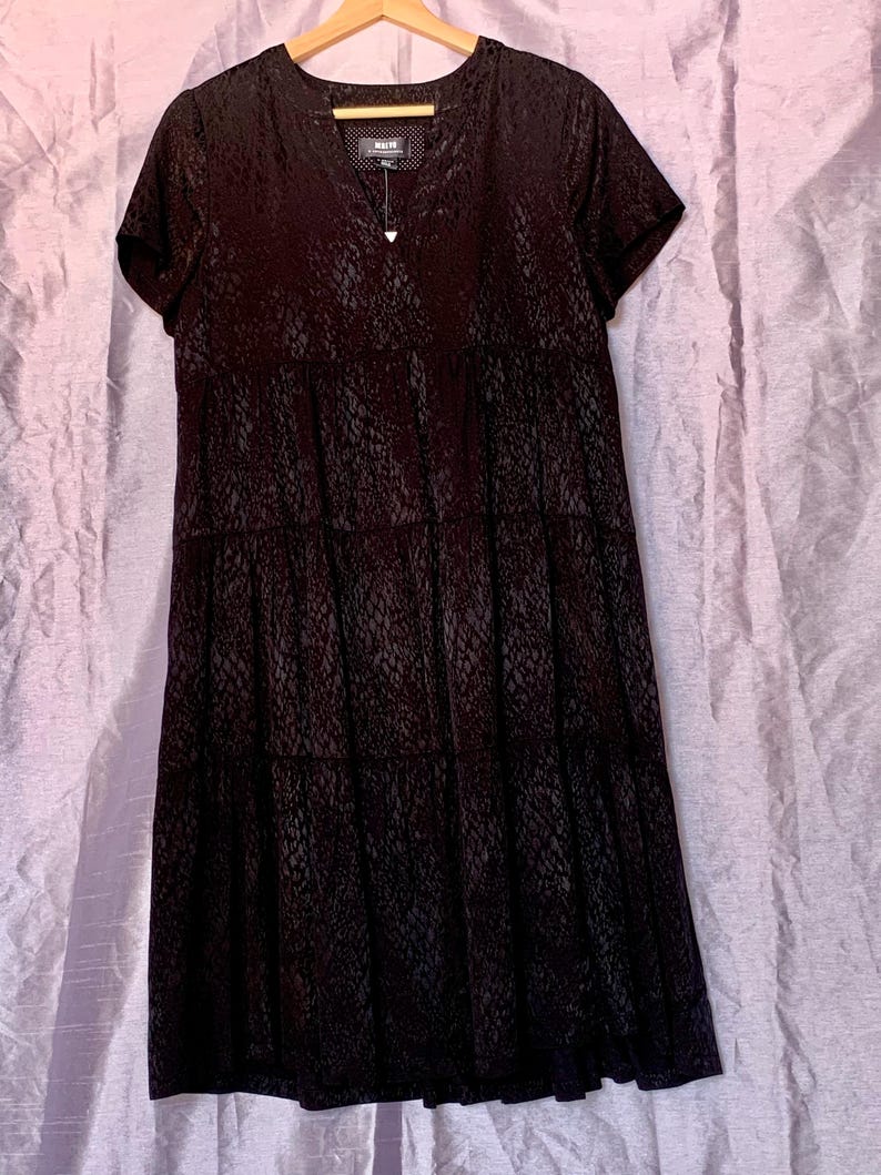 May include: A black, short-sleeved dress with a V-neck and a tiered skirt. The dress features a subtle, textured pattern. It hangs on a wooden hanger against a light purple backdrop.
