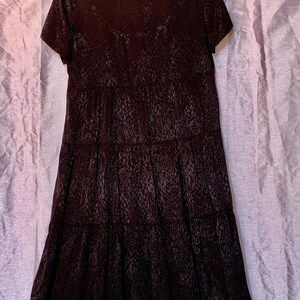 May include: A dark brown dress with short sleeves and a textured pattern resembling reptile skin. The dress has a tiered skirt design. It is hanging on a wooden hanger against a light purple backdrop. The dress is a stylish garment.