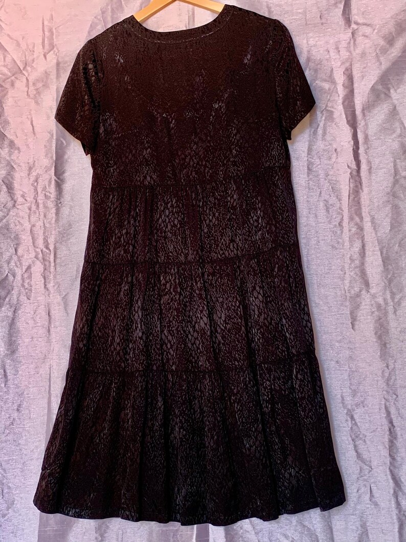 May include: A dark brown dress with short sleeves and a textured pattern resembling reptile skin. The dress has a tiered skirt design. It is hanging on a wooden hanger against a light purple backdrop. The dress is a stylish garment.