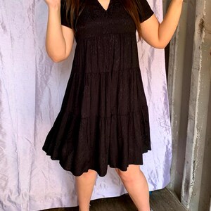 May include: A black tiered dress with short sleeves and a V-neck. The dress features a textured pattern on the bodice and a flowing skirt. The model is wearing black heels. The background includes a purple curtain and a grey wall.