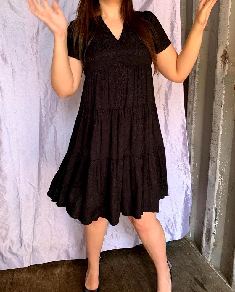 May include: A black tiered dress with short sleeves and a V-neck. The dress features a textured pattern on the bodice and a flowing skirt. The model is wearing black heels. The background includes a purple curtain and a grey wall.