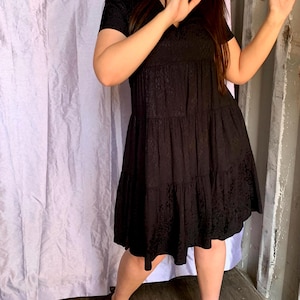 May include: A black, short-sleeved dress with a V-neck and tiered skirt. The dress has a subtle, textured pattern throughout. The model is wearing black heels. The background includes a white curtain and a wooden wall.