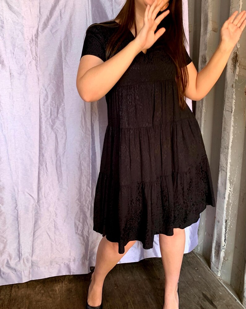 May include: A black, short-sleeved dress with a V-neck and tiered skirt. The dress has a subtle, textured pattern throughout. The model is wearing black heels. The background includes a white curtain and a wooden wall.