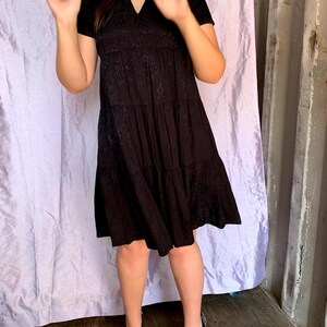 May include: A black tiered dress with a V-neck and short sleeves. The dress has a textured pattern on the bodice and skirt. The person is wearing black heels. The background is a light purple curtain and a grey wall.