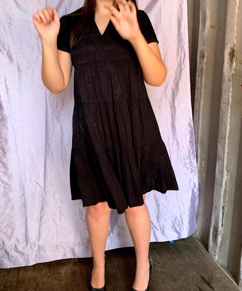May include: A black tiered dress with a V-neck and short sleeves. The dress has a textured pattern on the bodice and skirt. The person is wearing black heels. The background is a light purple curtain and a grey wall.