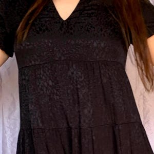 May include: A black dress featuring a V-neck and short sleeves. The dress has a textured pattern and a tiered skirt. The dress is made of a soft, flowing fabric, suitable for casual or semi-formal wear.