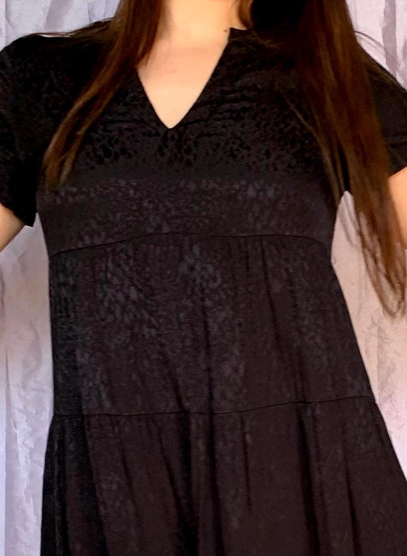 May include: A black dress featuring a V-neck and short sleeves. The dress has a textured pattern and a tiered skirt. The dress is made of a soft, flowing fabric, suitable for casual or semi-formal wear.