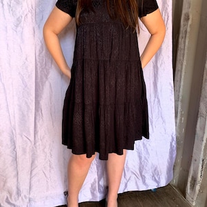 May include: A black, short-sleeved dress with a tiered skirt and a subtle, textured pattern. The dress has a V-neck and is paired with black heels. The dress is knee-length and has a relaxed fit.