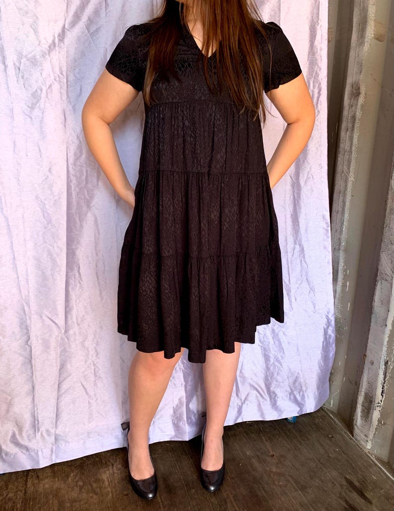 May include: A black, short-sleeved dress with a tiered skirt and a subtle, textured pattern. The dress has a V-neck and is paired with black heels. The dress is knee-length and has a relaxed fit.