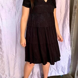 May include: A black, short-sleeved dress with a V-neck and tiered skirt. The dress has a subtle pattern and is paired with black heels. The dress is knee-length and appears to be made of a lightweight fabric.