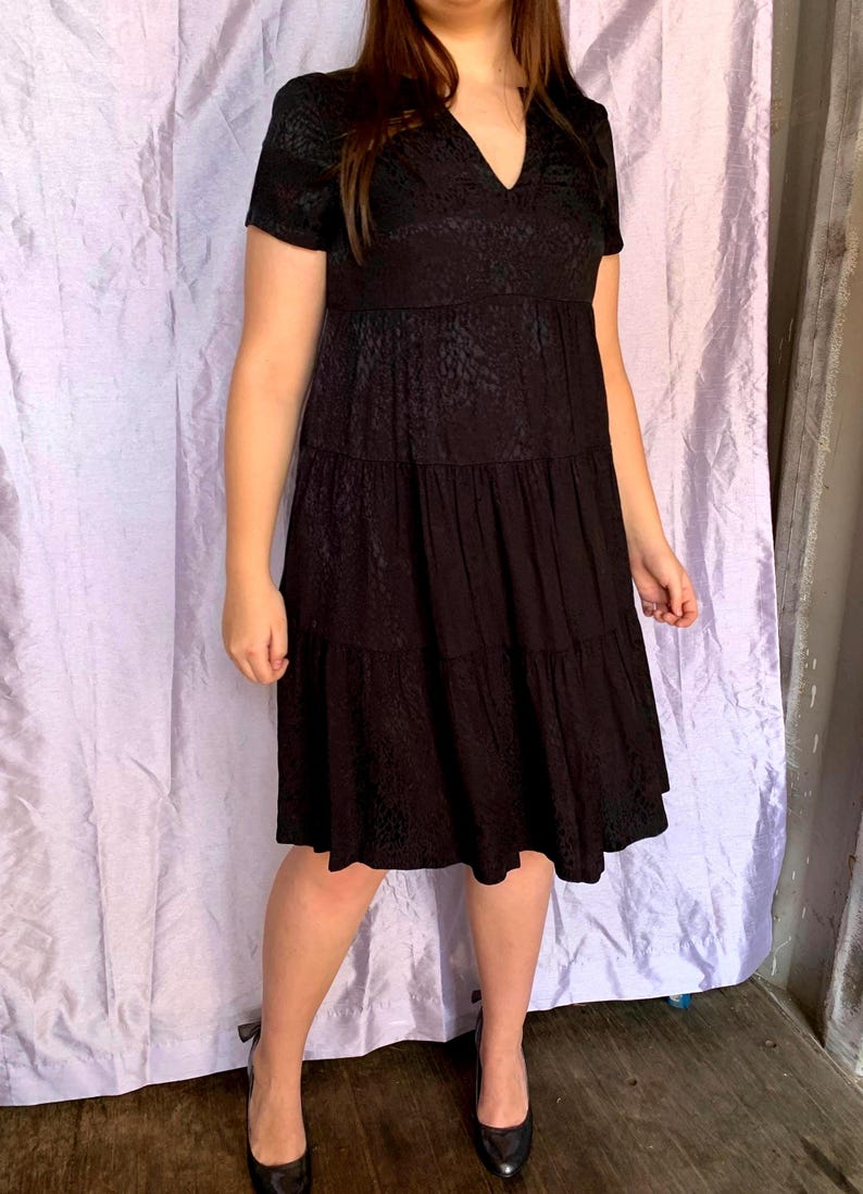 May include: A black, short-sleeved dress with a V-neck and tiered skirt. The dress has a subtle pattern and is paired with black heels. The dress is knee-length and appears to be made of a lightweight fabric.