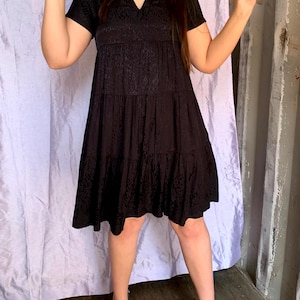 May include: Black tiered dress with a V-neck and short sleeves. The dress features a subtle pattern and falls to the knees. The model is wearing black heels. The background includes a light purple curtain and a grey wall.