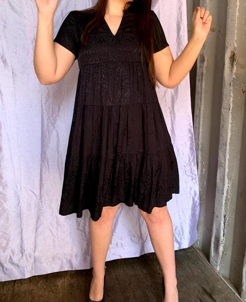 May include: Black tiered dress with a V-neck and short sleeves. The dress features a subtle pattern and falls to the knees. The model is wearing black heels. The background includes a light purple curtain and a grey wall.