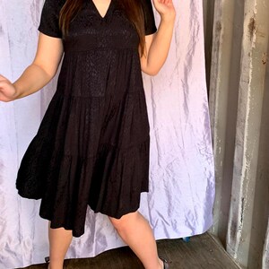 May include: A black, short-sleeved dress with a V-neck and tiered skirt. The dress features a subtle, textured pattern. The model wears black flats. The background includes a purple curtain and a grey wall.