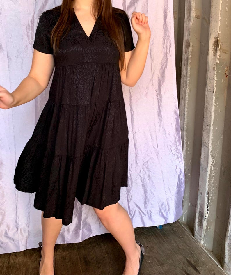 May include: A black, short-sleeved dress with a V-neck and tiered skirt. The dress features a subtle, textured pattern. The model wears black flats. The background includes a purple curtain and a grey wall.